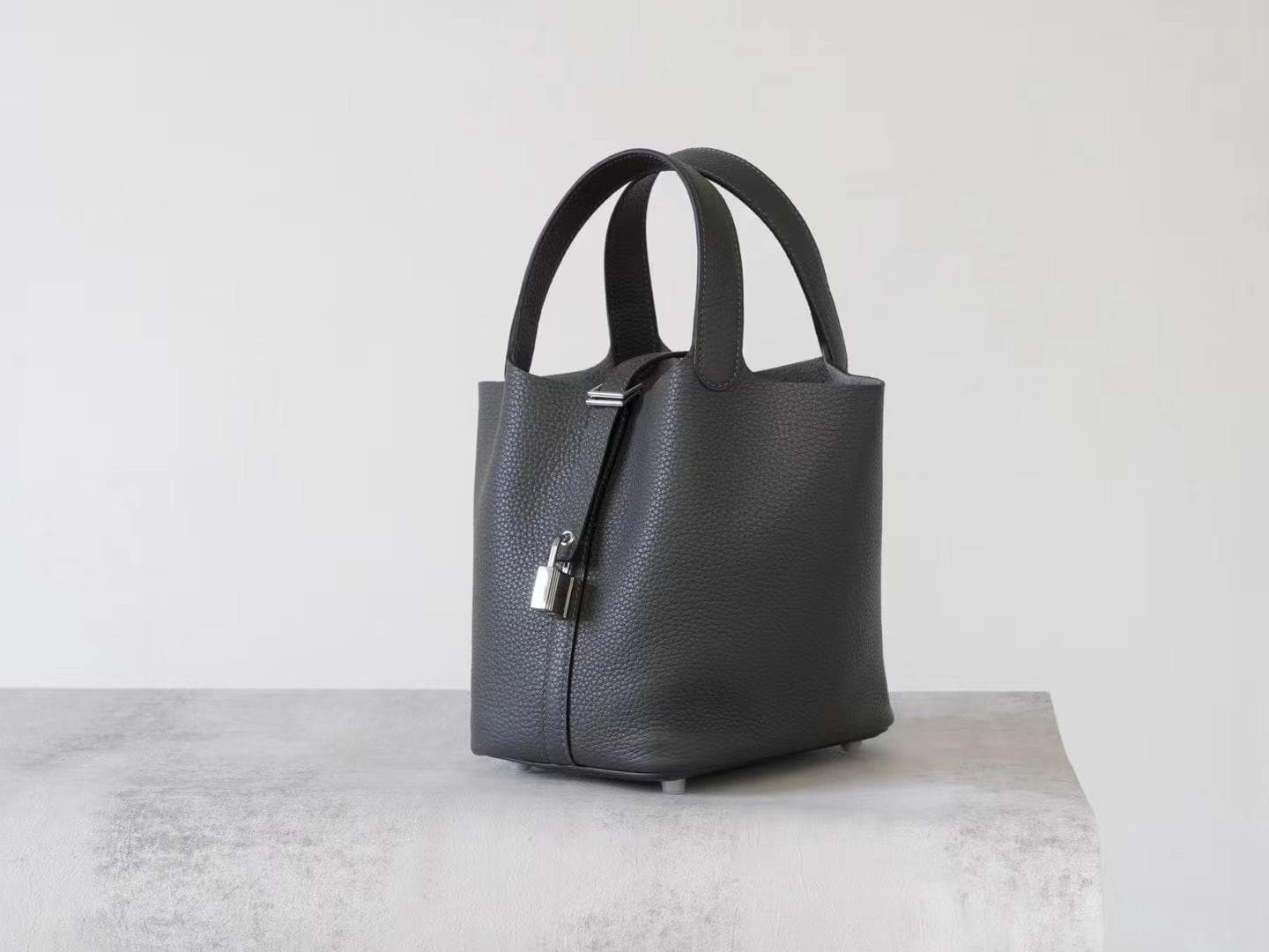 Linjin Luxury black pebbled leather bucket bag with dual top handles, center strap buckle closure, and silver padlock with key charm pendant, displayed on a woven rattan stool - 2